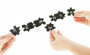 Three hands putting pieces into a puzzle that has the word 'TEAMWORK' written in chalk